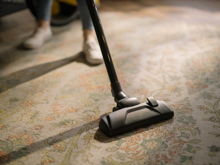 Close-up of a black vacuum cleaner nozzle being used to clean a patterned area rug in a domestic setting, with the person operating it partially visible in the background wearing beige shoes and dark 