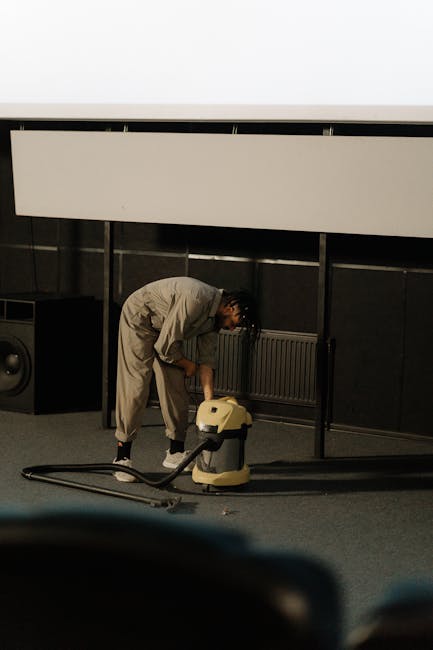 A person wearing a light-colored jumpsuit is engaged in surface cleaning using a vacuum cleaner on a dark, carpeted floor in an indoor space resembling a conference room or auditorium. The individual 