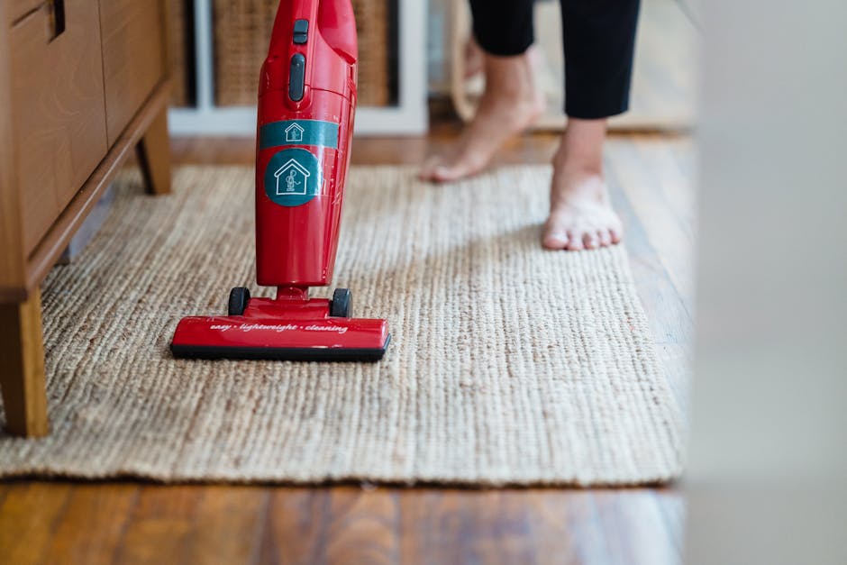A person wearing a light-colored jumpsuit is engaged in surface cleaning using a vacuum cleaner on a dark, carpeted floor in an indoor space resembling a conference room or auditorium. The individual 